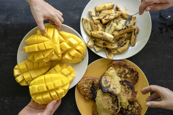 Bananpannkakor, stekt bananer, gul mango och människor händer. Smaskiga dessert, närbild, uppifrån — Stockfoto