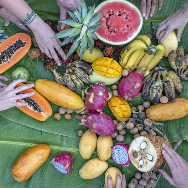 Tropiska frukter sortiment på en grön bananblad och människor händer. Smaskiga dessert, närbild. Mango, papaya, pitahaya och händer, uppifrån — Stockfoto