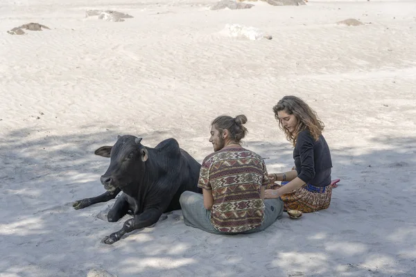 Молодой парень с девушкой отдыхает в жаркий солнечный день на берегу реки Ганга со священной коровой в городе Ришикеш, Индия — стоковое фото