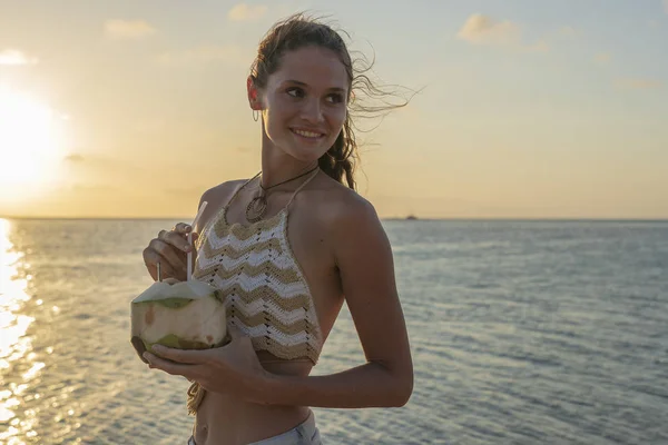Joven chica de belleza bebiendo coco en la playa tropical cerca del agua de mar en la isla paradisíaca al atardecer. Concepto de verano. Viaje de vacaciones . — Foto de Stock