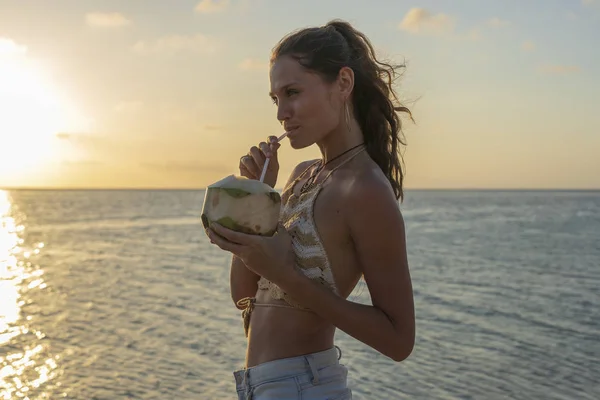 Joven chica de belleza bebiendo coco en la playa tropical cerca del agua de mar en la isla paradisíaca al atardecer. Concepto de verano. Viaje de vacaciones . — Foto de Stock