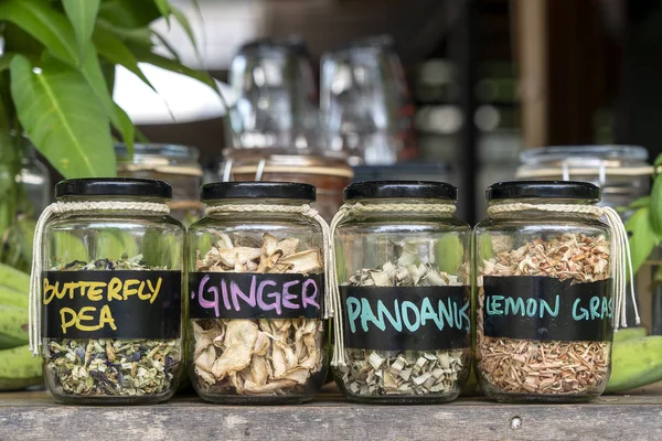 Assortment of dried spices in glass bottles on wooden background. Dry ginger, pandanus, lemon grass and butterfly pea in glass jars, closeup — Stock Photo, Image