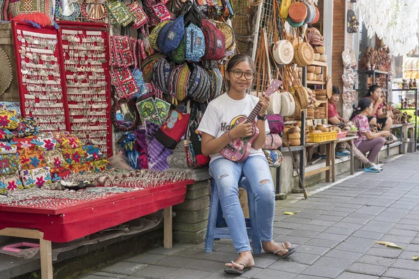 Joven chica indonesia toca la guitarra y ofrece comprar de sus recuerdos a los turistas en un mercado callejero en la ciudad de Ubud, isla Bali, Indonesia —  Fotos de Stock