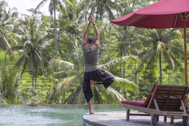 Tropikal yüzme havuzunda yakınındaki ağaç pozisyonda meditasyon ve Yoga yapıyor adam Bali, Endonezya Adası
