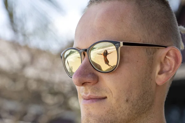 Retrato de un joven en gafas de sol en las que la silueta de una niña se refleja en la playa tropical cerca del mar — Foto de Stock