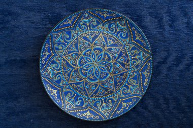 Mavi ve altın renklerde dekoratif seramik plaka, mavi kumaş arka planda boyalı plaka, closeup. Akrilik boyalar ile boyanmış dekoratif porselen plaka, el işi