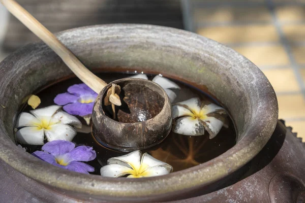 Scoop kokosnoot shell en een aardewerk jar gevuld met water samen met bloemen. Thailand — Stockfoto