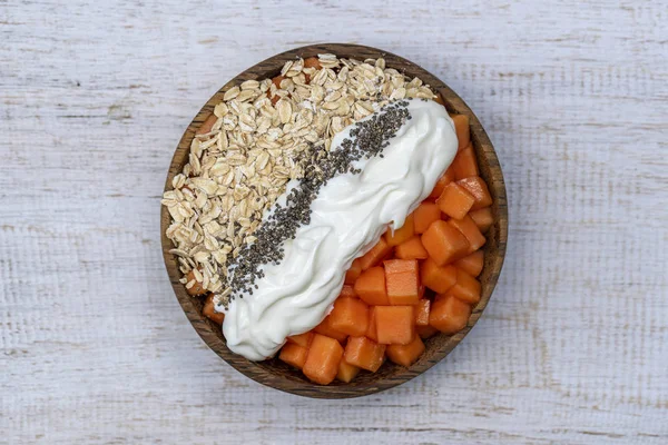 Slices of sweet papaya with oat flakes, chia seeds and white yogurt in coconut bowl on white wooden background, close up — Stock Photo, Image