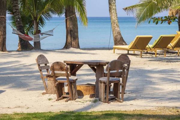 Tropisk sandstrand med träbord och stolar, schäslonger, hängmatta och palmträd mot bakgrund av det blå havet, Thailand. Rese-och natur koncept. — Stockfoto