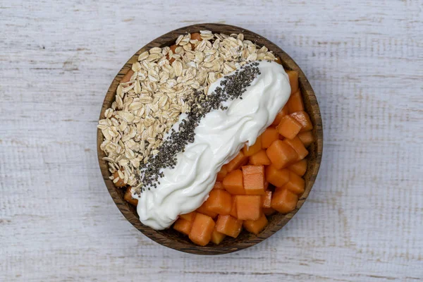 Slices of sweet papaya with oat flakes, chia seeds and white yogurt in coconut bowl on white wooden background, close up — Stock Photo, Image