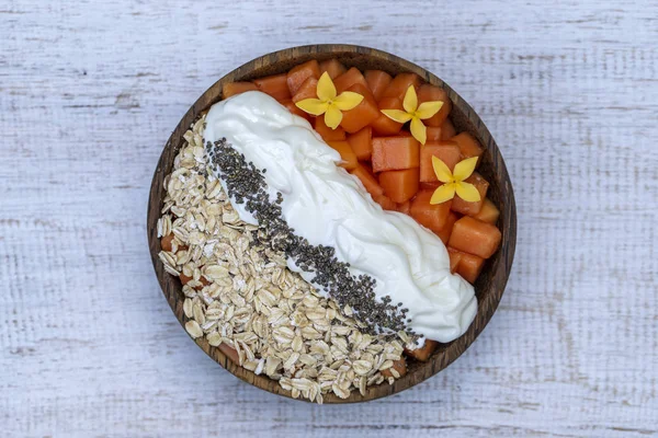 Scheiben süße Papaya mit Haferflocken, Chiasamen und weißem Joghurt in Kokosnussschale auf weißem Holzgrund, Nahaufnahme — Stockfoto