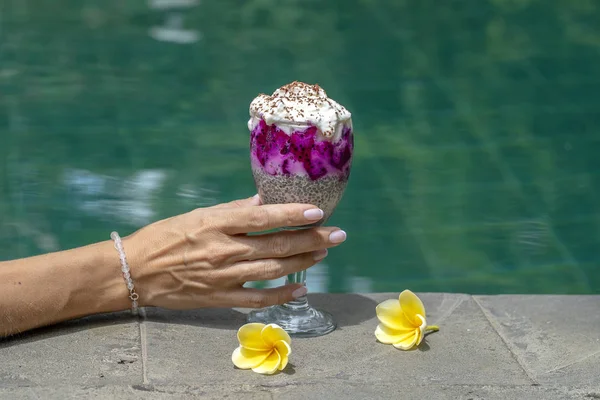 Dívka ruka drží sklenici s hia semena pudink na pozadí vody v bazénu. Chia semínka pudink s červený drak ovoce a bílého jogurtu ve sklenici k snídani — Stock fotografie