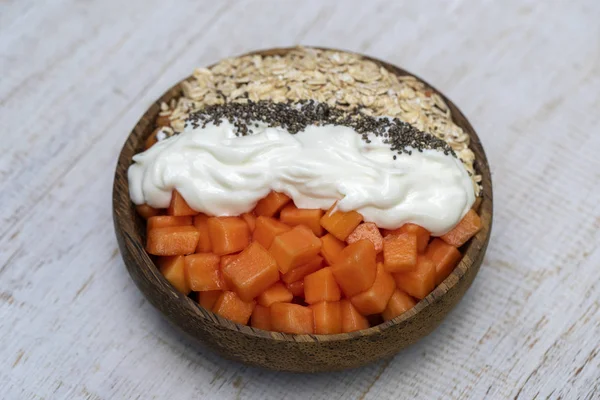 Fatias de mamão doce com flocos de aveia, sementes de chia e iogurte branco em tigela de coco sobre fundo de madeira branca, close up — Fotografia de Stock