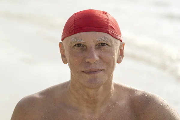 Oudere man in een rode zwemmen hoed op het strand in de buurt van het zeewater — Stockfoto