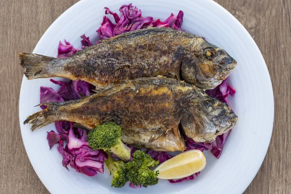 Две жареные морские рыбы подаются на тарелке с красной капустой, зеленой брокколи и лимоном. Закрыть — стоковое фото