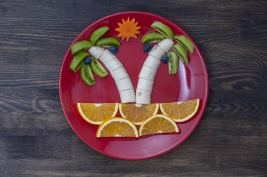 Kivi, muz, üzüm, havuç ve portakallı yaratıcı meyve tatlısı. Çocuk maması kavramı. Eğlenceli yemek. Ahşap zemin üzerinde kırmızı tabakta meyvelerden yapılmış palmiye ağaçları.