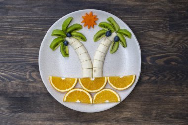 Kivi, muz, üzüm, havuç ve portakallı yaratıcı meyve tatlısı. Çocuk maması kavramı. Eğlenceli yemek. Ahşap zemin üzerinde beyaz tabakta meyvelerden yapılmış palmiye ağaçları.