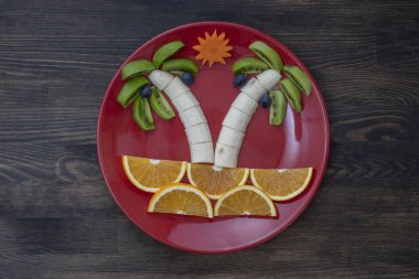 Kivi, muz, üzüm, havuç ve portakallı yaratıcı meyve tatlısı. Çocuk maması kavramı. Eğlenceli yemek. Ahşap zemin üzerinde kırmızı tabakta meyvelerden yapılmış palmiye ağaçları.