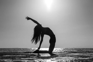 Silhouette of woman standing at yoga pose on the tropical beach during sunset. Caucasian girl practicing yoga near sea water. Black and white