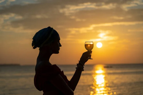 热带海滩上 一个可爱的女人在海浪边 面对美丽的落日 举着一杯白葡萄酒 女孩正在喝着一杯葡萄酒欣赏日落 休闲和旅行的概念 — 图库照片