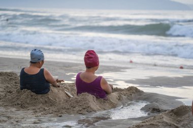Danang, Vietnam - june 15, 2020 : Two vietnames women taking a bath of beach sand for therapy near sea water in the morning on the city Danang, Vietnam. Local people relax on the beach clipart