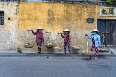 Hoi An, Vietnam - 16 Mart 2020: Vietnam 'daki Hoi An, Vietnam' daki eski sarı duvarın arkasında elinde meyve sepeti olan hasır şapkalı Vietnamlı kadınlar