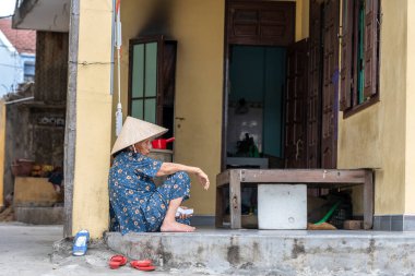 Hoi An, Vietnam - 16 Mart 2020: Hasır şapkalı yaşlı Vietnamlı kadın Hoi An, Vietnam 'daki evinin yakınındaki bir sokakta oturuyor
