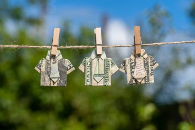Asılı yeşil doğa arka planı ve mavi gökyüzü üzerine dolar banknotundan yapılmış Origami gömleği. İpe asılmış dolar banknotları. Kapatın. Dolar Tişörtü