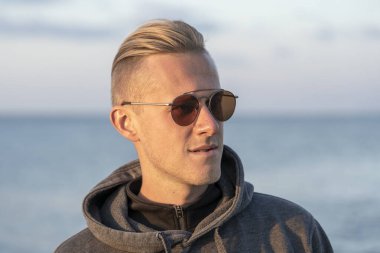 Güneş gözlüklü bir gencin portresi. Sahilde, sonbaharda, deniz kenarında dinleniyor. Yaklaş.