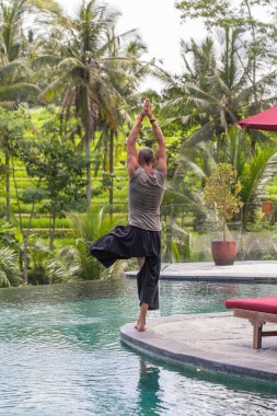 Adam yoga yapıyor ve Endonezya 'nın Bali adasındaki tropikal plajda havuz kenarında ağaç pozisyonunda meditasyon yapıyor. Sağlıklı yaşam tarzı kavramı