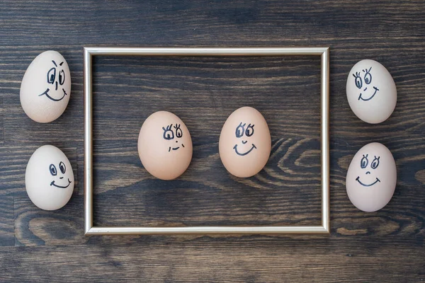 Picture golden frame and many funny eggs smiling on dark wooden wall background, close up. Eggs family emotion face portrait. Concept funny food