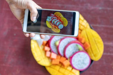 Kadın eli tropikal meyvelerin fotoğrafını çekiyor. Kahvaltıda çekilmiş akıllı telefon fotoğrafı, sosyal medya ya da blog için. Tatlı mango, papaya, pitahaya. Çiğ vejetaryen sağlıklı yiyecekler