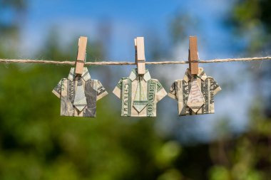 Asılı yeşil doğa arka planı ve mavi gökyüzü üzerine dolar banknotundan yapılmış Origami gömleği. İpe asılmış dolar banknotları. Kapatın. Dolar Tişörtü