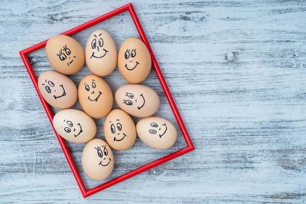 Picture red frame and many funny eggs smiling on white wooden wall background, close up. Eggs family emotion face portrait. Concept funny food. Copy space