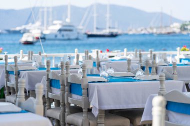 Masa örtüsü ve çatalı olan beyaz yemek masaları, bıçak, şarap bardağı, tabak ve restoranda mavi peçete. Akşam yemeği için gümüş takımlar ve peçeteyle güzel bir masa seti, Bodrum, Turkey. Deniz kenarındaki sahil kafe.
