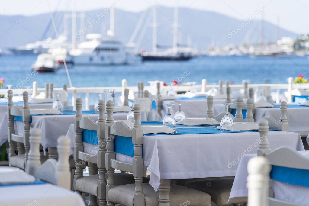 White dining tables with tablecloth and fork, knife, wine glass, plate and blue napkin in restaurant. Nice table set with arranged silverware and napkin for dinner, Bodrum, Turkey. Beach cafe near sea