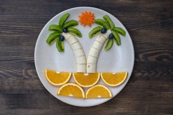 Kivi, muz, üzüm, havuç ve portakallı yaratıcı meyve tatlısı. Çocuk maması kavramı. Çocuklar için eğlenceli ve sağlıklı meyve salatası. Ahşap zemin üzerinde beyaz tabakta meyvelerden yapılmış hurma ağaçları.