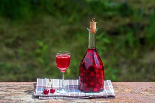 赤チェリーの自家製チンキ ベリーアルコール飲料のコンセプト ガラス瓶に熟したチェリーから作られた自家製の赤ワインと緑の庭の背景にある木製のテーブルの上のワイングラス ウクライナ — ストック写真