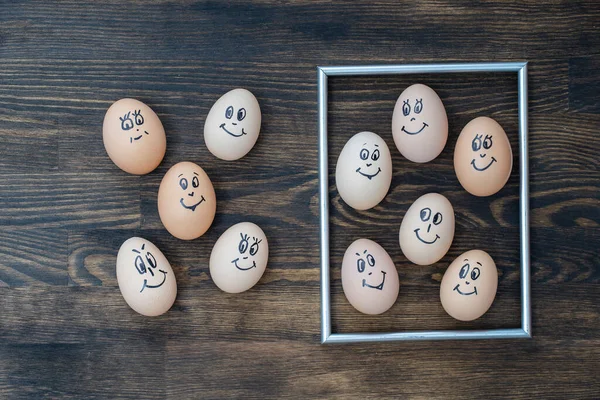 Picture golden frame and many funny eggs smiling on dark wooden wall background, close up. Eggs family emotion face portrait. Concept funny food