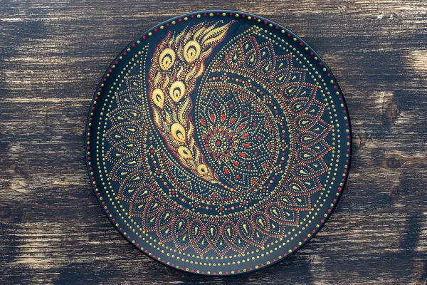 Decorative ceramic plate with black, red and golden colors, painted plate on wooden background, close up, top view. Decorative porcelain plate painted with acrylic paints, handwork, dot painting