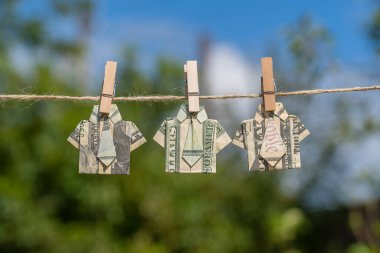 Asılı yeşil doğa arka planı ve mavi gökyüzü üzerine dolar banknotundan yapılmış Origami gömleği. İpe asılmış dolar banknotları. Kapatın. Dolar Tişörtü