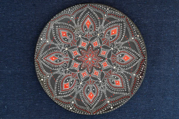 Decorative ceramic plate with red and silver colors, painted plate on background of blue fabric, closeup, top view. Decorative porcelain plate painted with acrylic paints, handwork, dot painting