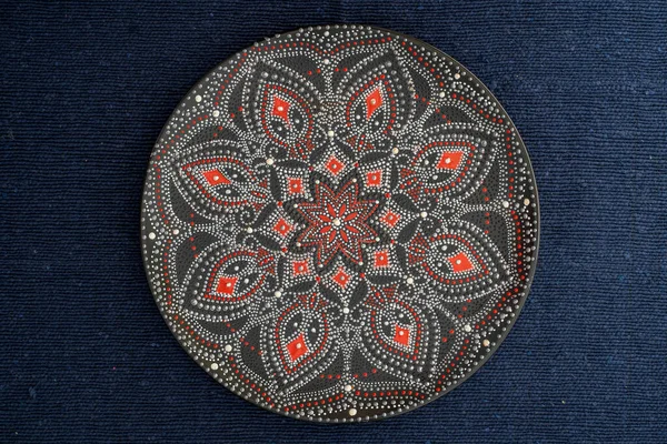 Decorative ceramic plate with black, red and golden colors, painted plate on background of fabric, closeup, top view. Decorative porcelain plate painted with acrylic paints, handwork, dot painting