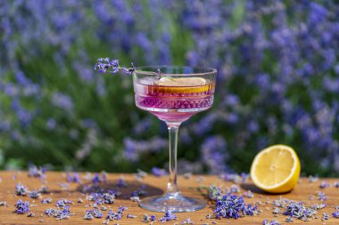 Yeşil bir yaz bahçesine karşı ahşap masada zarif kristal bardakta servis edilen parlak kokteyl ya da limonata. Eflatun çiçekli mor renkli içecek ve bir dilim limon.