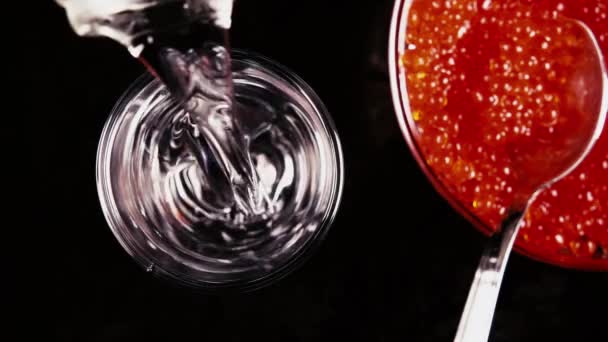Slow motion giet de wodka in een glas op een zwarte lijst met rode kaviaar bovenaanzicht — Stockvideo