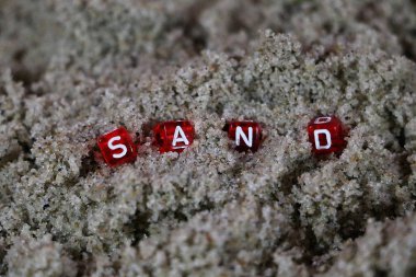 Kumsalda beyaz harflerle küçük kırmızı zarları kapatın ve kum kelimesini yazın.
