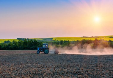 the tractor pulls the plow in the field on beautiful sunset or sunrise clipart