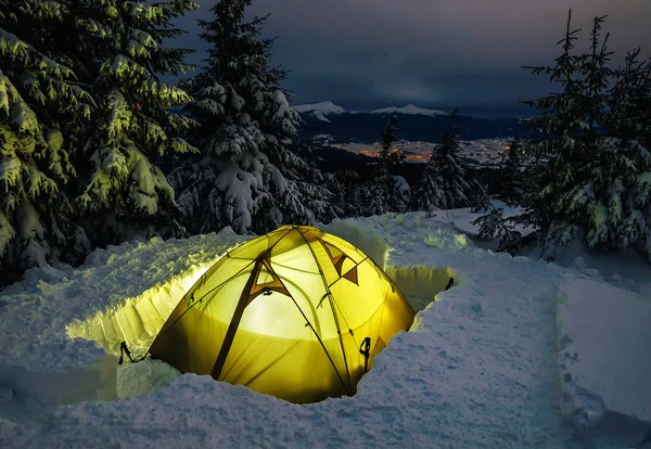 Winter camping, night, shining green tent on snow, clouds, forrest. Night shot, long exposure, sleeping on snow in the outdoors.