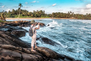 Şapkalı kız Sri Lanka 'da günbatımında akıllı telefondan okyanusun fotoğraflarını çekiyor.