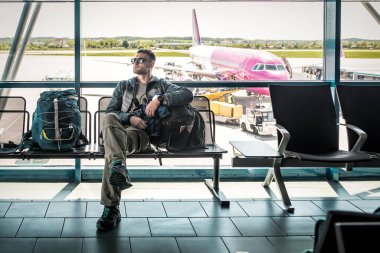 Güneş gözlüklü sırt çantalı bir adam havaalanı terminalinde uçmayı bekliyor.